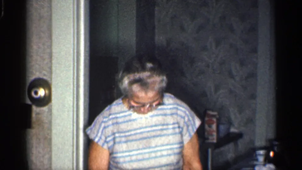 In a 1959 California home interior, an older woman in glasses and a striped top looks down while standing near a doorway.