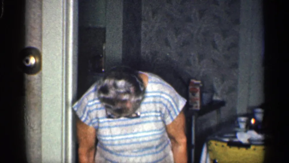 An older woman in a striped top leans forward near a doorway, with wallpaper and household items visible behind her.