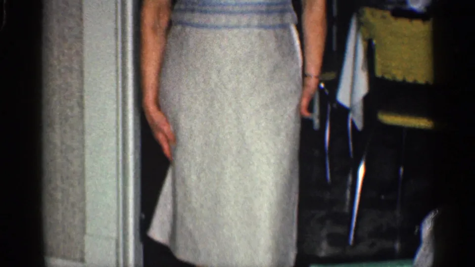 The woman stands near the doorway with her light skirt visible, framed by dim mid-century home furnishings.