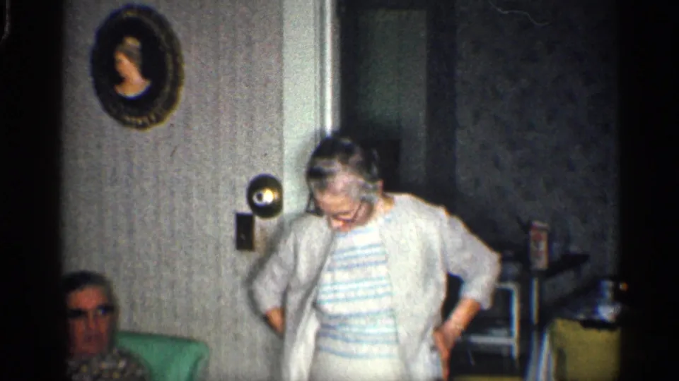 In the home interior, the older woman adjusts her clothing as another adult face appears partly visible at the lower left.