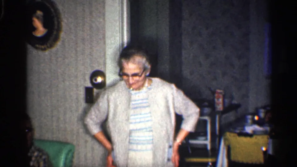 The woman faces the camera in the 1959 California interior, wearing glasses and a light cardigan over a striped dress near a wall portrait.