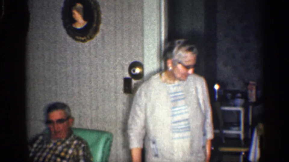 The woman turns beside a seated man in glasses and a plaid shirt, with a mint-green armchair and mid-century room decor visible.