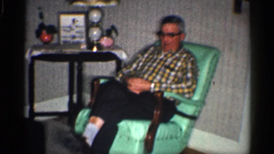 A man in glasses and a plaid shirt sits in a mint-green armchair in the California home room, near a small table and framed decor.