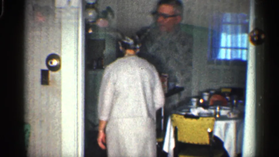 1959 California home-movie frame showing a woman entering a private dining or kitchen area with a man near a table.