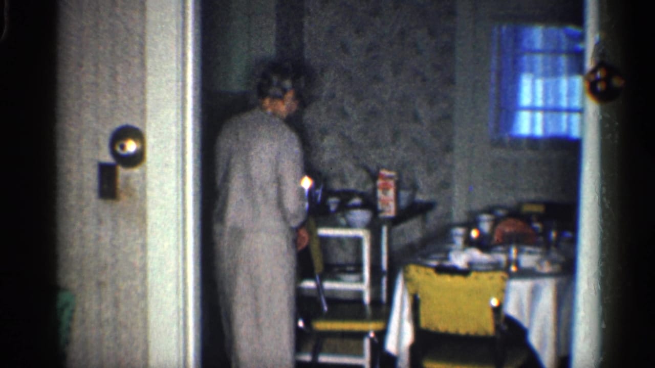 Woman in a gray dress stands beside a yellow chair near a table in a small room.