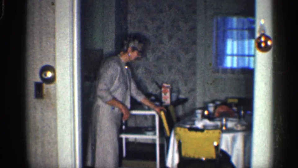 1959 California home-movie frame showing a woman adjusting or pushing a yellow chair beside a table.