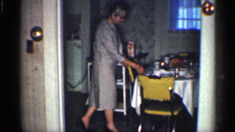 1959 California home-movie frame of a woman by a table and yellow chair inside a private kitchen or dining room.