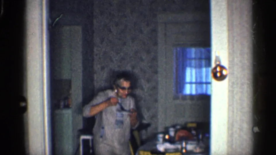 A woman in sunglasses appears to begin a playful gesture or dance in a 1959 California kitchen seen through a doorway.