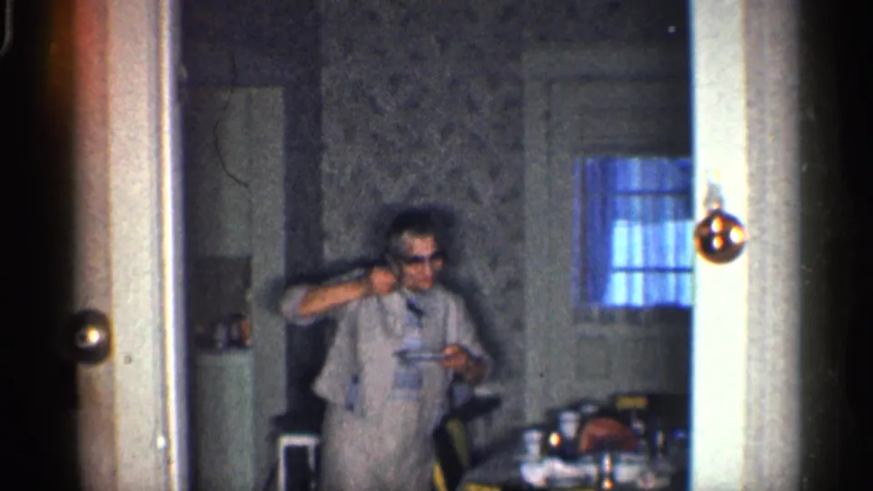 The woman faces the camera in the kitchen, holding a small object while framed by the doorway and patterned wallpaper.