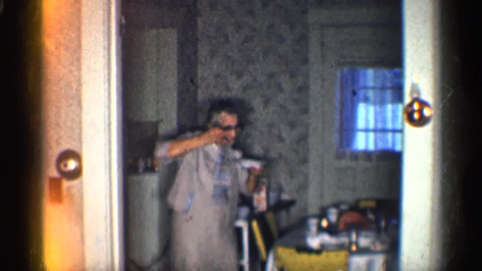 A woman in a light dress raises her arm as if dancing beside a kitchen table and yellow chair in the California home interior.