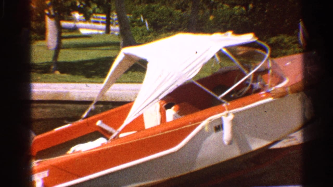 1959 California home-movie frame showing a red-and-white speedboat with a white canvas canopy near a grassy lakeside area.