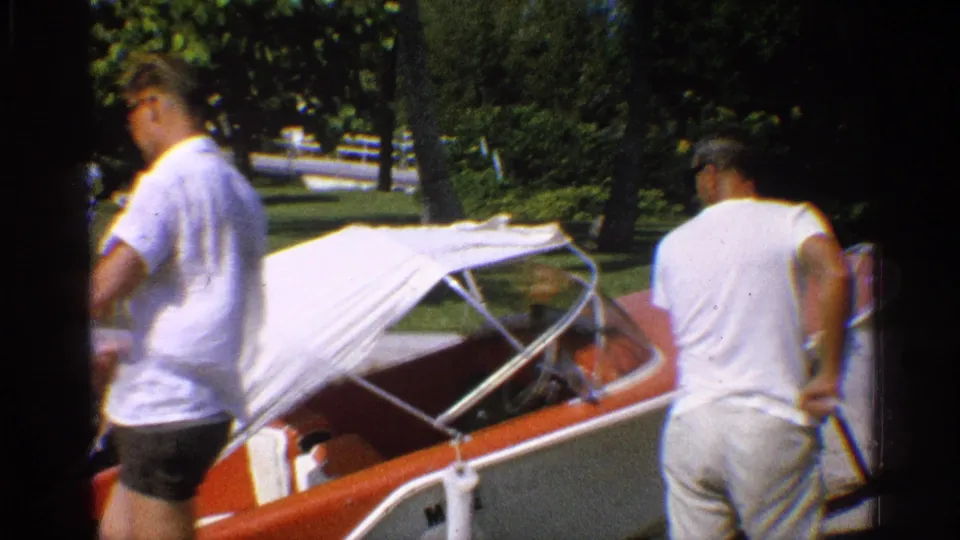 Two men in light casual clothing stand by the red-and-white speedboat as it is positioned near water in 1959 California footage.