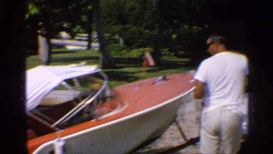 A man in a white shirt and light pants stands behind the boat while the trailer and bow are angled toward the launch area.
