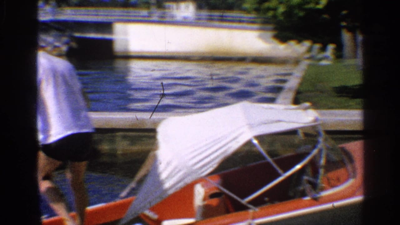 California 1959 home-movie frame showing a man near a red motorboat with a white canopy at a dock, with water and bridge structure behind.