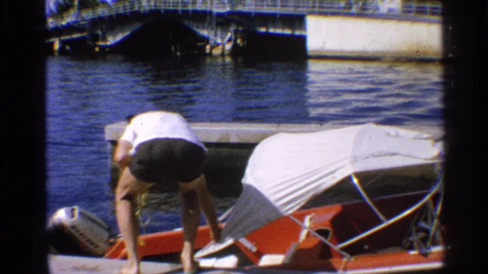 California 1959 frame of an adult man leaning over a red motorboat at the dock, appearing to adjust or secure the white canvas canopy.