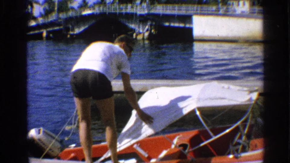 California 1959 8mm frame showing a man in a white shirt and dark shorts reaching across the white canopy of a red docked motorboat.