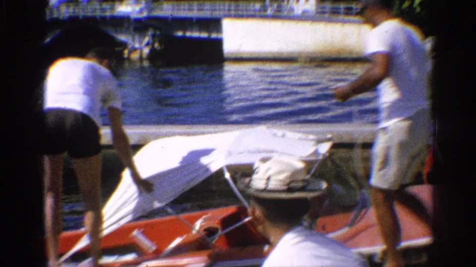 California 1959 frame showing two adult men by a red motorboat at a dock, one working over the white canopy with calm water in the background.
