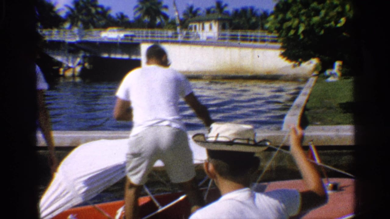Man in white shirt stands by a covered motorboat on a canal with bridge and palm trees behind him.