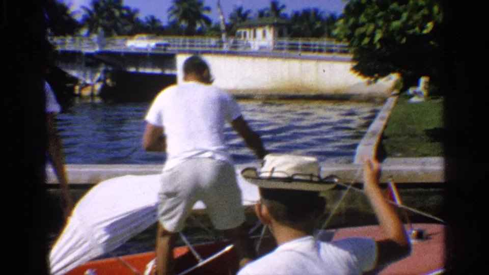 1959 California home-movie frame showing a man in a white shirt beside a covered motorboat on a canal, with a bridge and palms in the background.