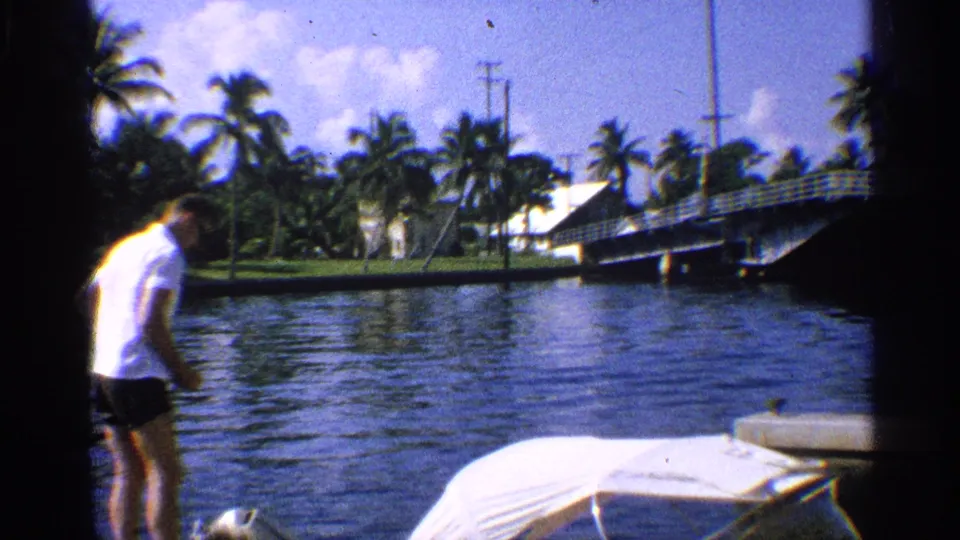 1959 California home-movie frame of a man at the canal edge, with a covered motorboat, palm trees, shoreline structures, and a bridge visible.