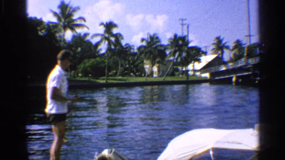 1959 California canal-side home-movie frame with a man in a white shirt beside the water, a covered boat in front, and palms across the shore.
