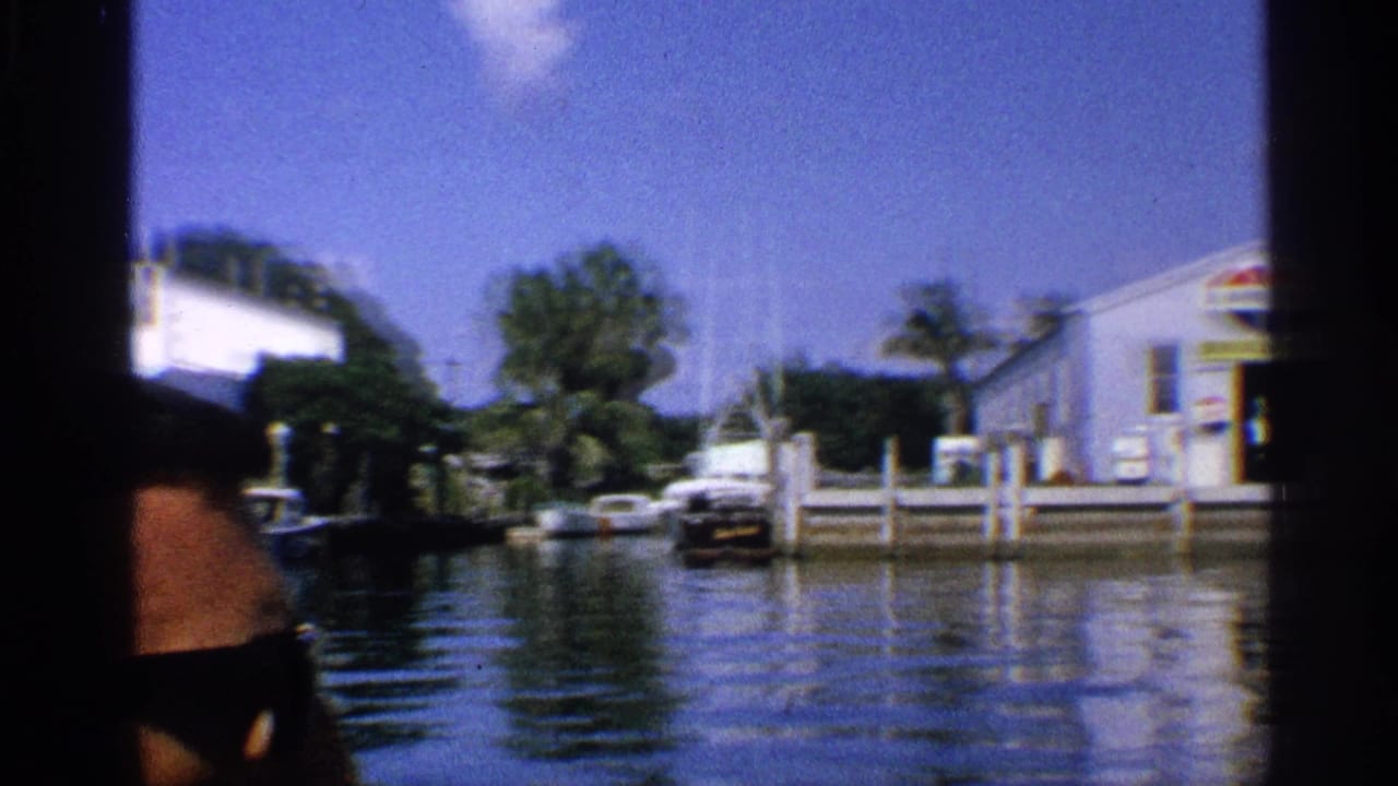 1959 California 8mm frame with a partly visible man in sunglasses looking toward a marina, boats, docks, and white buildings across the water.