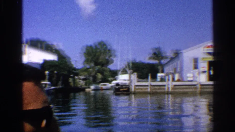 1959 California 8mm frame with a partly visible man in sunglasses looking toward a marina, boats, docks, and white buildings across the water.