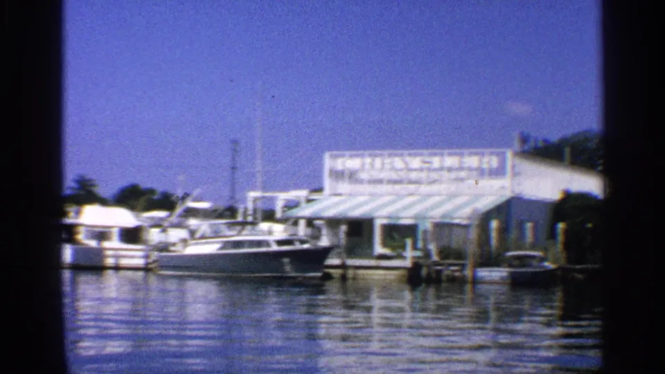 1959 California 8mm frame of docked pleasure boats, still marina water, a white waterfront building, and a striped awning under a bright sky.