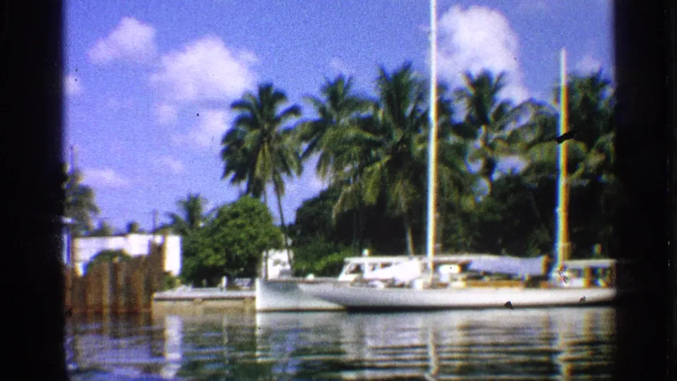 1959 California 8mm frame showing the sailboat from a slightly shifted angle, with palms, shore structures, and reflected water.