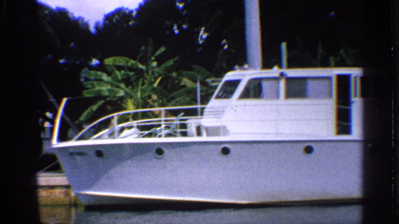 1959 California 8mm frame of a white cabin motorboat docked in a palm-lined waterway with dark foliage behind it.