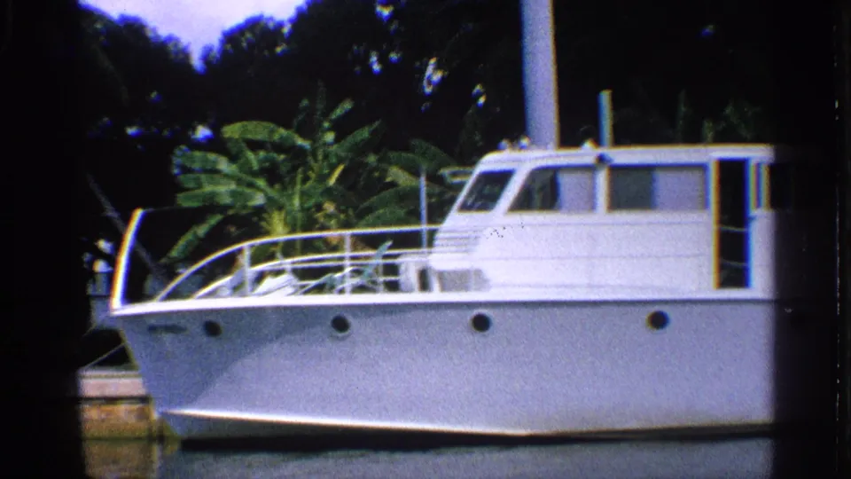 1959 California 8mm frame of a white cabin motorboat docked in a palm-lined waterway with dark foliage behind it.