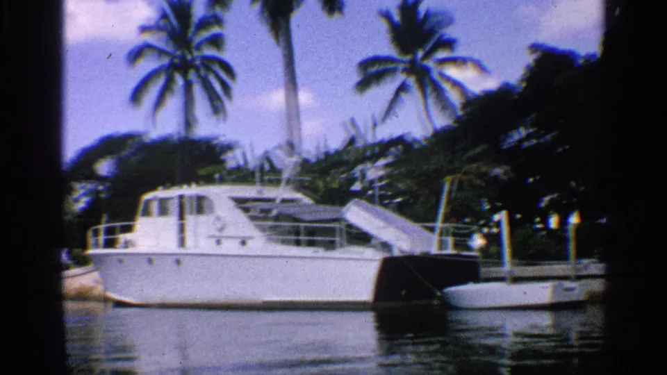 1959 California 8mm still of a white cabin motorboat moored near pilings, with tall palms and dense greenery behind the waterway.