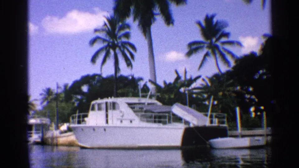 1959 California home-movie frame of a white cabin boat in a calm palm-lined waterway, with dock structures and smaller boats nearby.