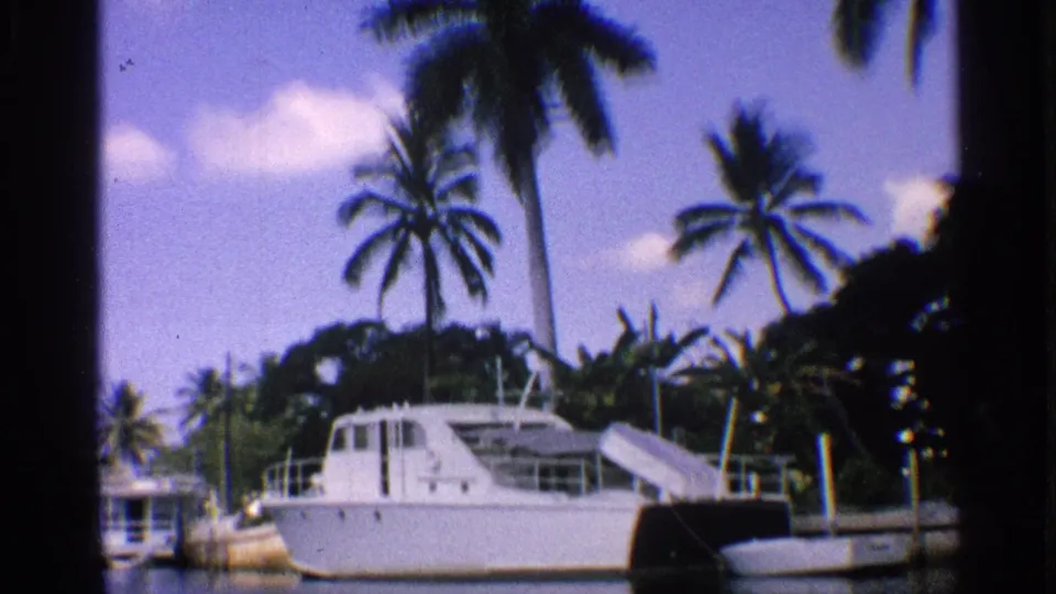 1959 California 8mm frame showing a white cabin boat at a dock with palm trees, calm water, and dark film-frame edges.