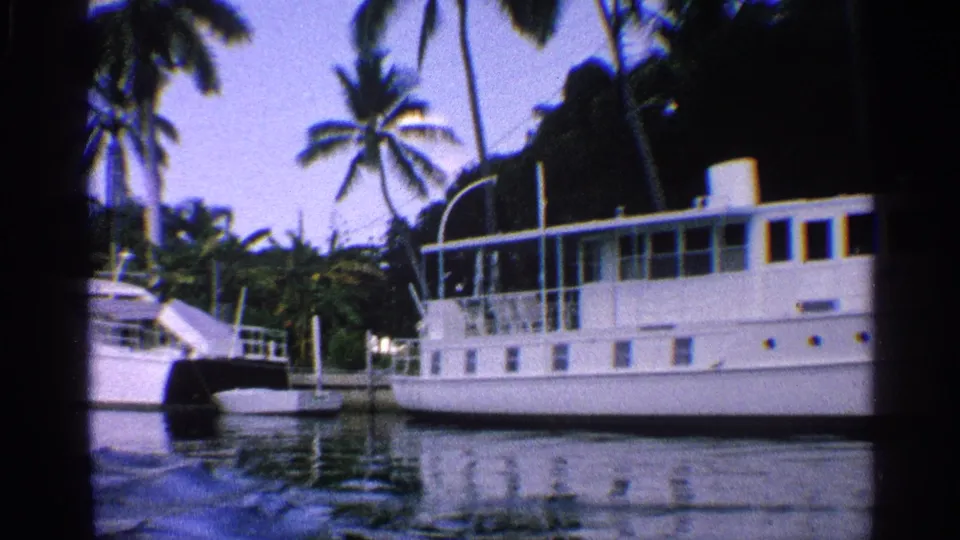 1959 California home-movie still of a large white waterfront boat, nearby dock structures, palms, and reflections on the canal.