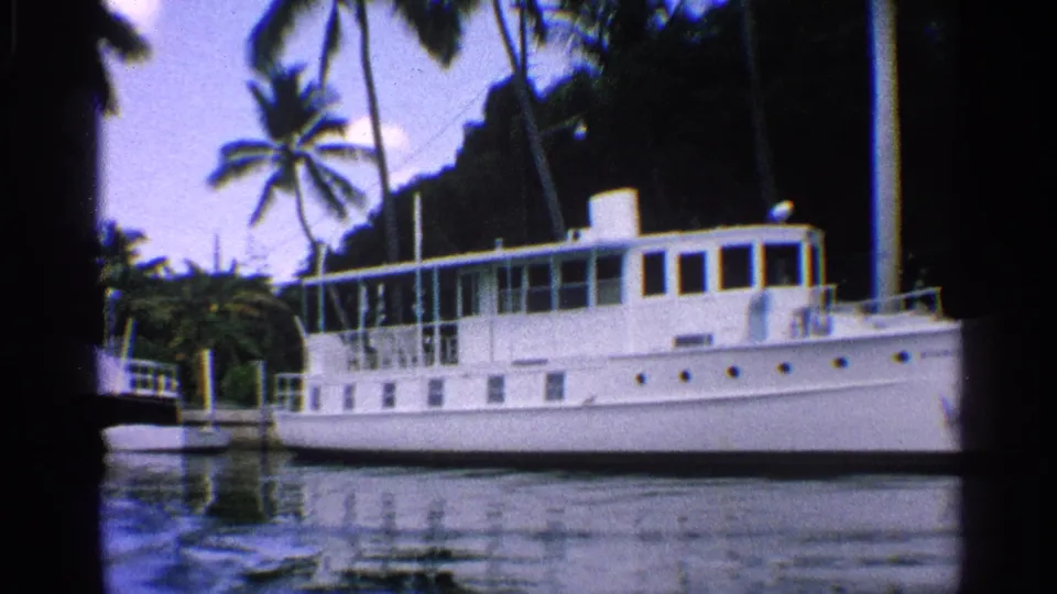 1959 California 8mm frame with a long white boat in profile, dock posts, palm trees, and rippled canal reflections.