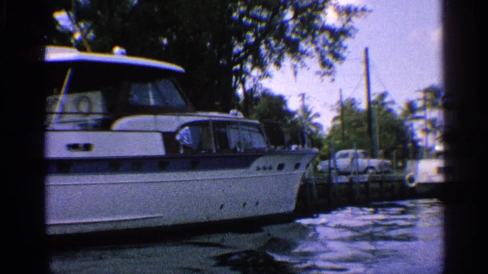 1959 California home-movie frame showing a white motorboat close to camera, dock pilings, palm trees, and shaded canal water.
