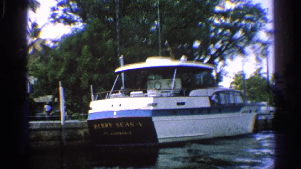 1959 California home-movie frame of MERRY SEAS-V from the stern, showing the yacht moored among canal pilings and greenery.