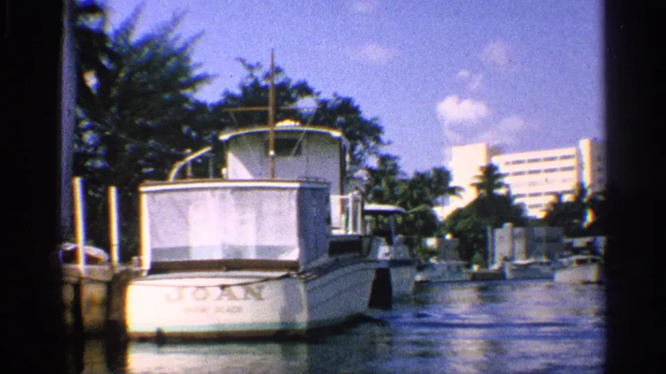 California 8mm frame showing the stern of a boat named JOAN in a canal, with palms and a distant white high-rise building.