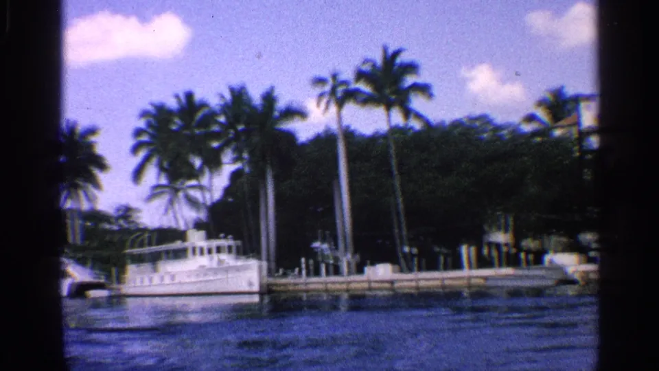 1959 California home-movie frame showing a white boat at the dock, tall palms, shaded shoreline trees, rippled canal water, and bright sky.