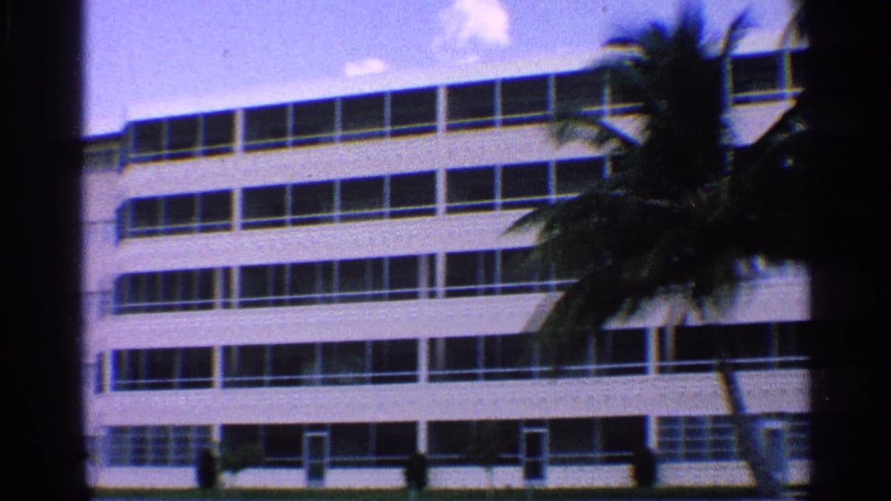 1959 California home-movie frame showing a white hotel-like building with horizontal window rows and palm trees in front.