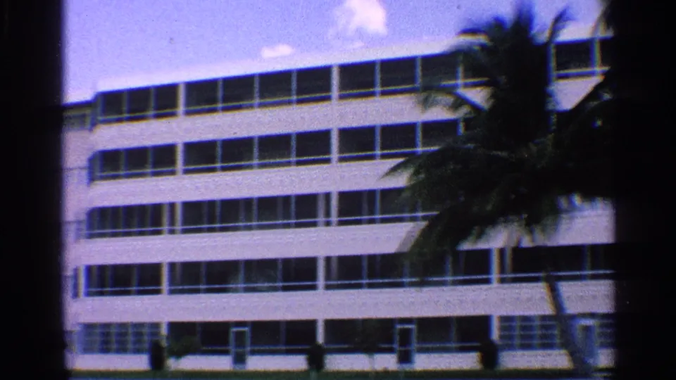 1959 California home-movie frame showing a white hotel-like building with horizontal window rows and palm trees in front.