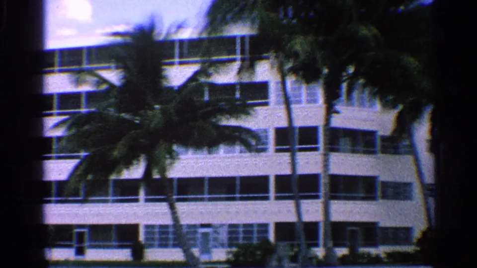 1959 California frame with palms crossing the facade of a white multi-story hotel or apartment-style building.
