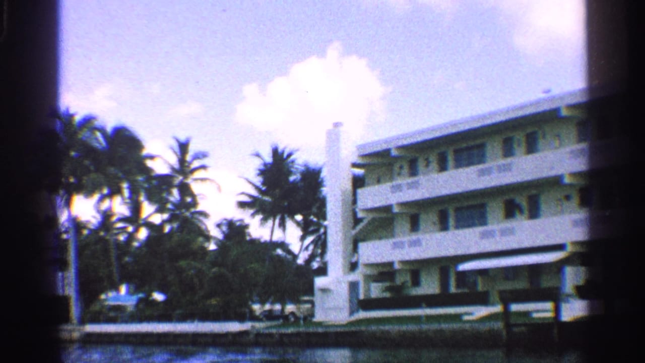 White multi-story hotel across water with palms and a bright sky.
