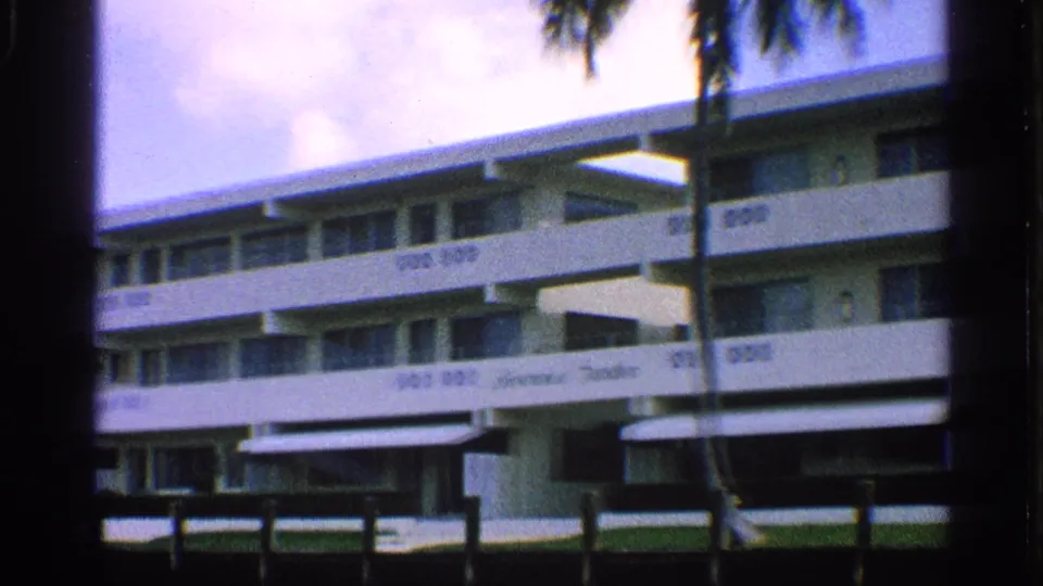 White mid-century hotel exterior with balconies, awnings, palm trunk, and waterfront foreground in California footage from 1959.