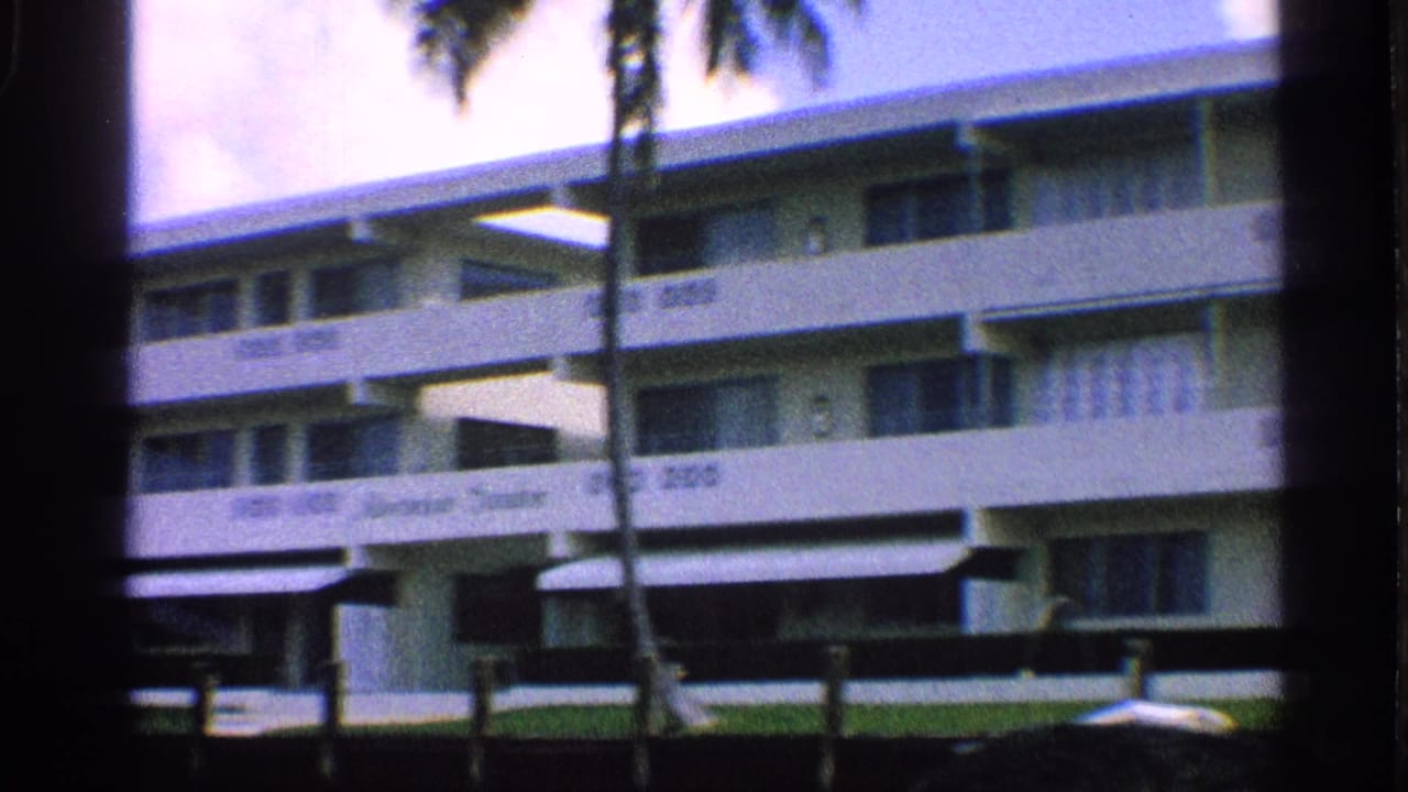 1959 California 8mm frame showing a white mid-century apartment building with balconies, palm trunks, lawn, and soft handheld focus.