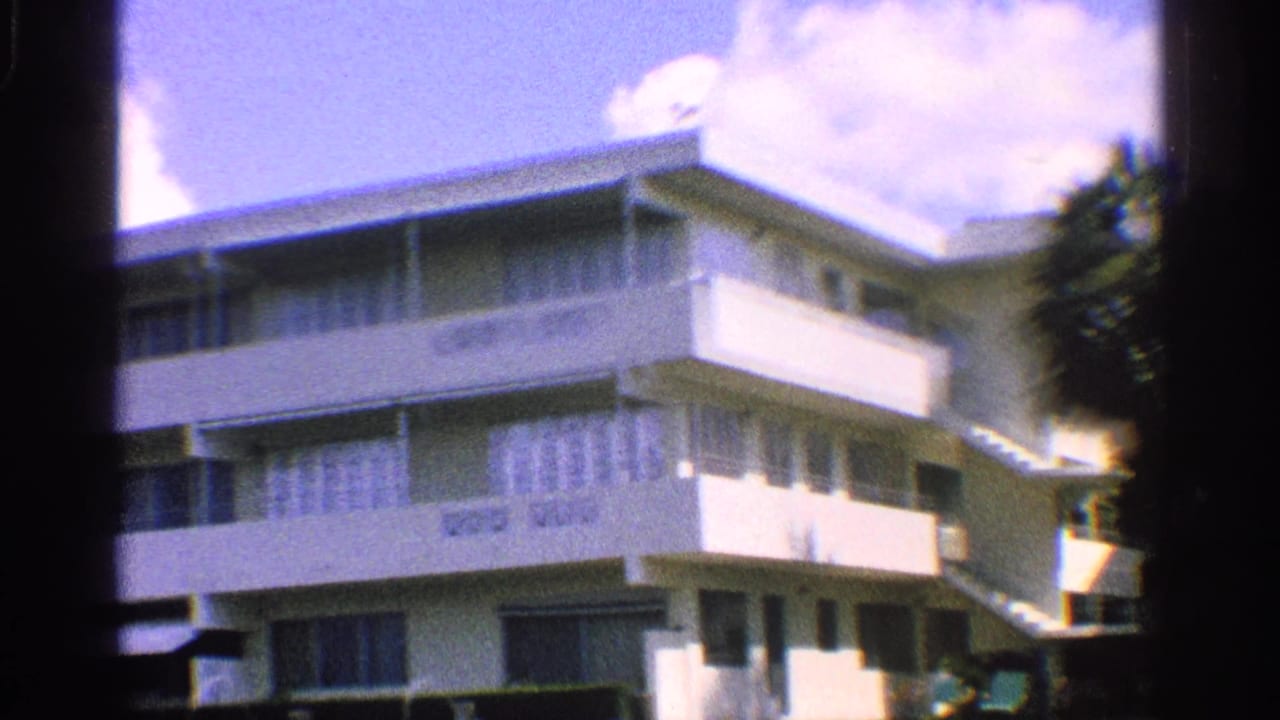 Corner of a white apartment building with balconies, windows, sky, and trees.