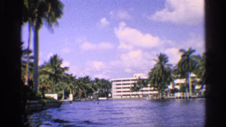 1959 California 8mm frame looking across water toward palm trees, white waterfront buildings, docks or seawalls, and scattered clouds.