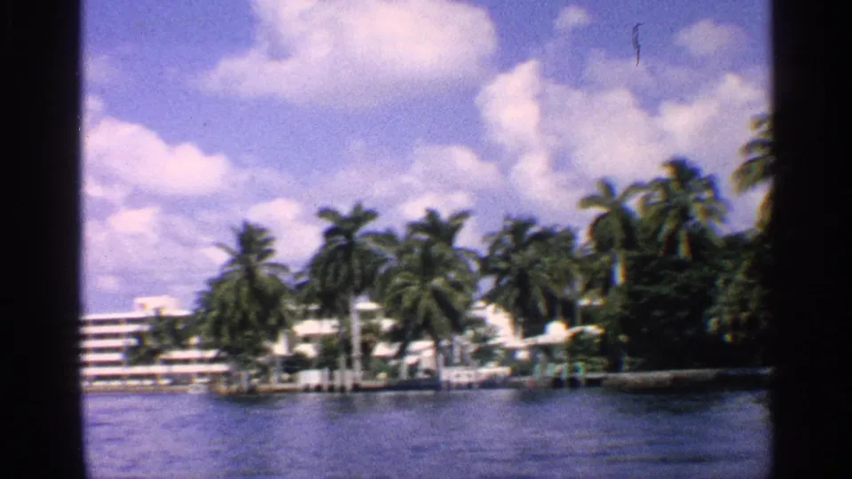 1959 California 8mm frame showing a palm-lined waterway with low white buildings, shoreline structures, and a bright partly cloudy sky.