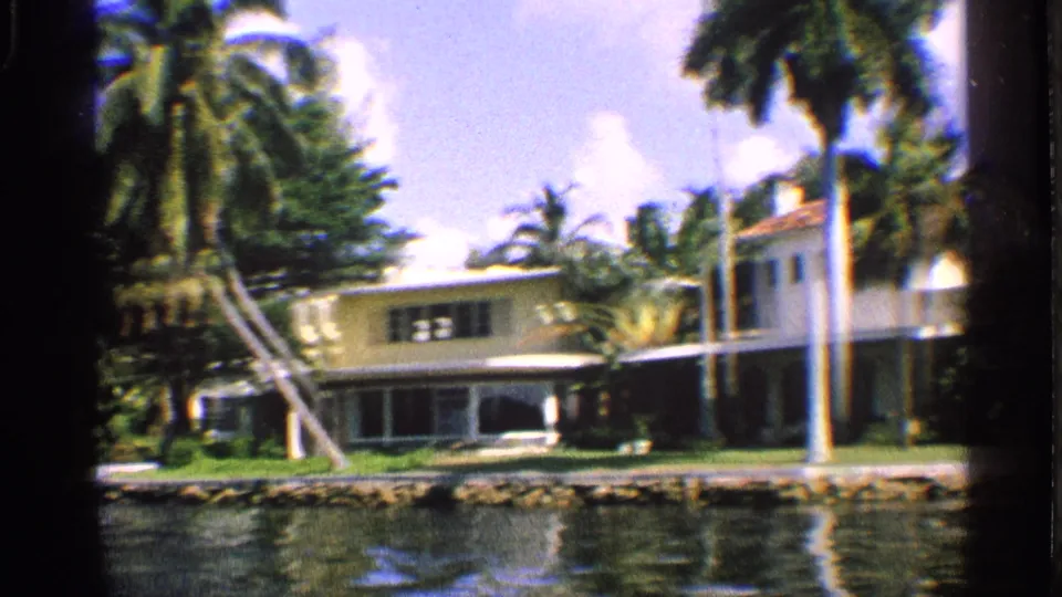 1959 California waterfront home with pale walls, tall palms, lawn, and rippling water in the foreground.